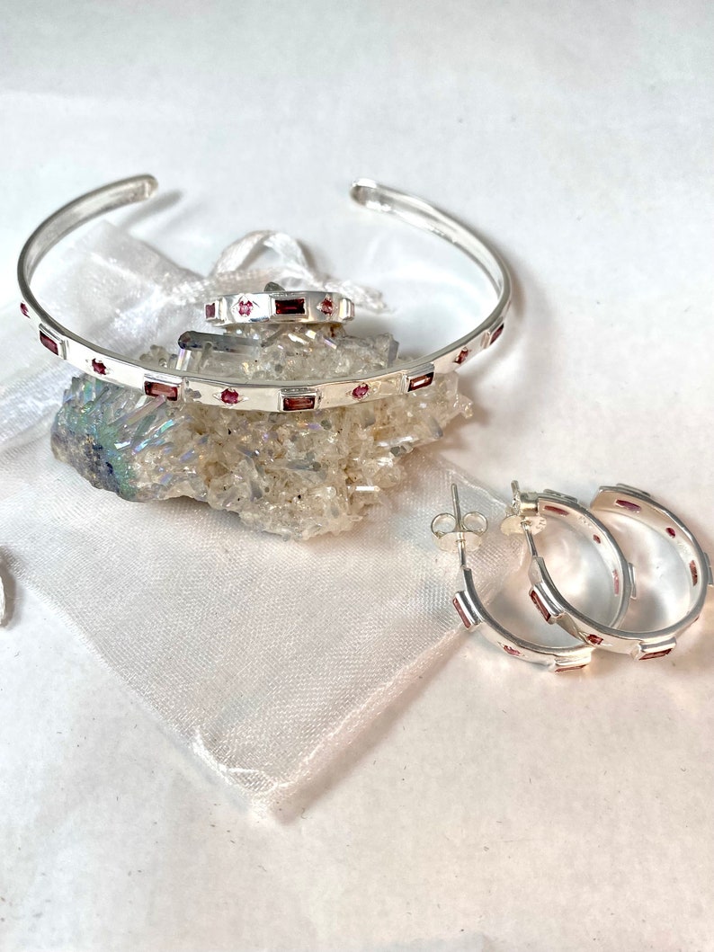 May include: A set of silver jewelry with pink gemstones. The set includes a bangle bracelet, a ring, and a pair of hoop earrings.