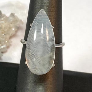 May include: A silver ring featuring a large, pear-shaped, translucent gemstone. The gemstone has a subtle, milky white and gray internal pattern. The ring is displayed on a black jewelry display stand, showcasing its elegant design.