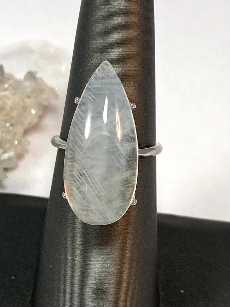 May include: A silver ring featuring a large, pear-shaped, translucent gemstone. The gemstone has a subtle, milky white and gray internal pattern. The ring is displayed on a black jewelry display stand, showcasing its elegant design.