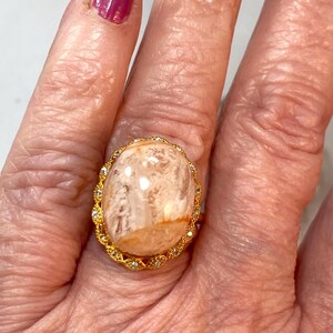 May include: A gold-toned ring featuring a large, oval-shaped, light-colored stone with a marbled appearance. The stone is surrounded by a delicate gold filigree setting, accented with small, clear gemstones. The ring is worn on a finger.