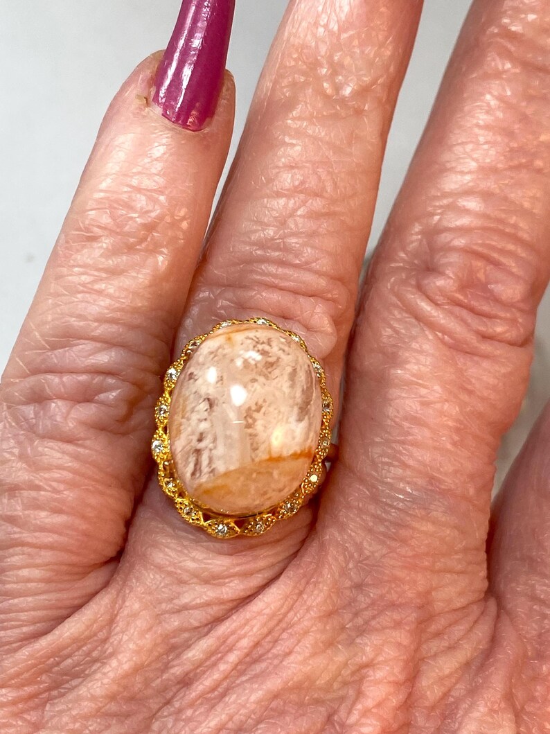 May include: A gold-toned ring featuring a large, oval-shaped, light-colored stone with a marbled appearance. The stone is surrounded by a delicate gold filigree setting, accented with small, clear gemstones. The ring is worn on a finger.