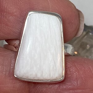 May include: A white rectangular ring with a silver band. The ring is made of a smooth, white material with a textured surface.