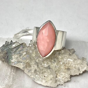 Pode incluir: Um anel de prata com uma gema de opala rosa engastada em forma de marquise. O anel est&aacute; sobre um fundo branco.