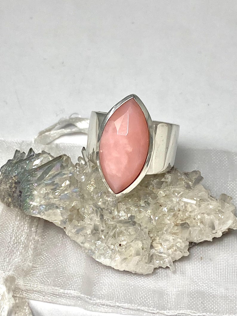 Pode incluir: Um anel de prata com uma gema de opala rosa engastada em forma de marquise. O anel est&aacute; sobre um fundo branco.
