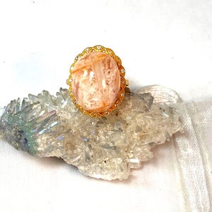 May include: A gold-toned ring featuring a large, oval-shaped, peach-colored gemstone. The gemstone is set in a decorative, filigree-style setting with small, clear accent stones. The ring is displayed on a cluster of clear quartz crystals.
