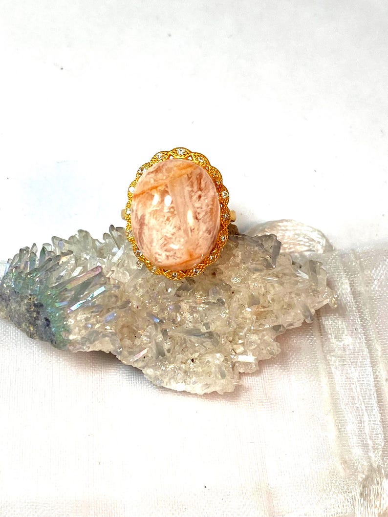 May include: A gold-toned ring featuring a large, oval-shaped, peach-colored gemstone. The gemstone is set in a decorative, filigree-style setting with small, clear accent stones. The ring is displayed on a cluster of clear quartz crystals.