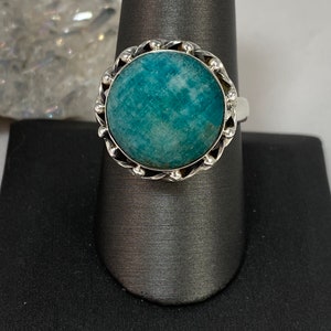 K&ouml;nnte beinhalten: Ein silberner Ring mit einem runden, blaugr&uuml;nen Edelstein, der in einer dekorativen silbernen Fassung sitzt. Der Ring befindet sich auf einem grauen Ringst&auml;nder.