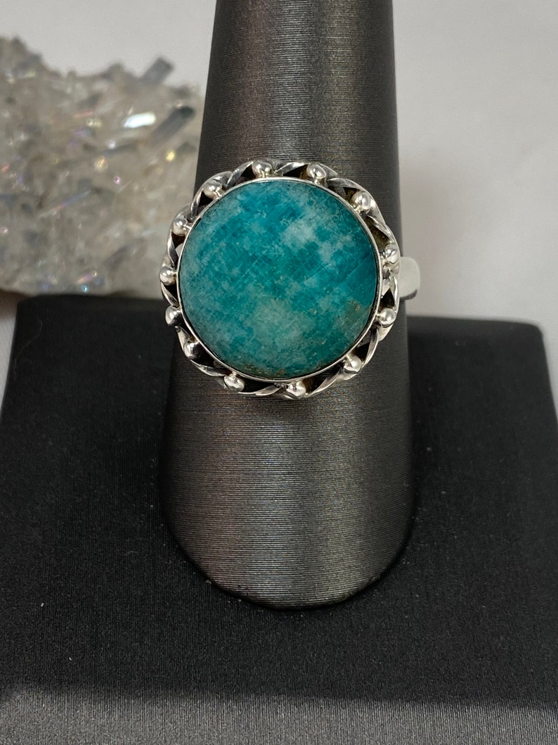 K&ouml;nnte beinhalten: Ein silberner Ring mit einem runden, blaugr&uuml;nen Edelstein, der in einer dekorativen silbernen Fassung sitzt. Der Ring befindet sich auf einem grauen Ringst&auml;nder.
