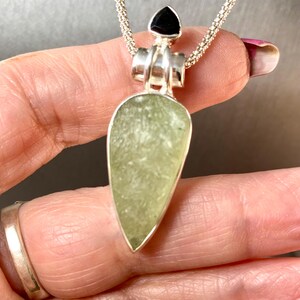 May include: A silver chain necklace with a pendant featuring a light green teardrop-shaped stone and a black triangular stone at the top.