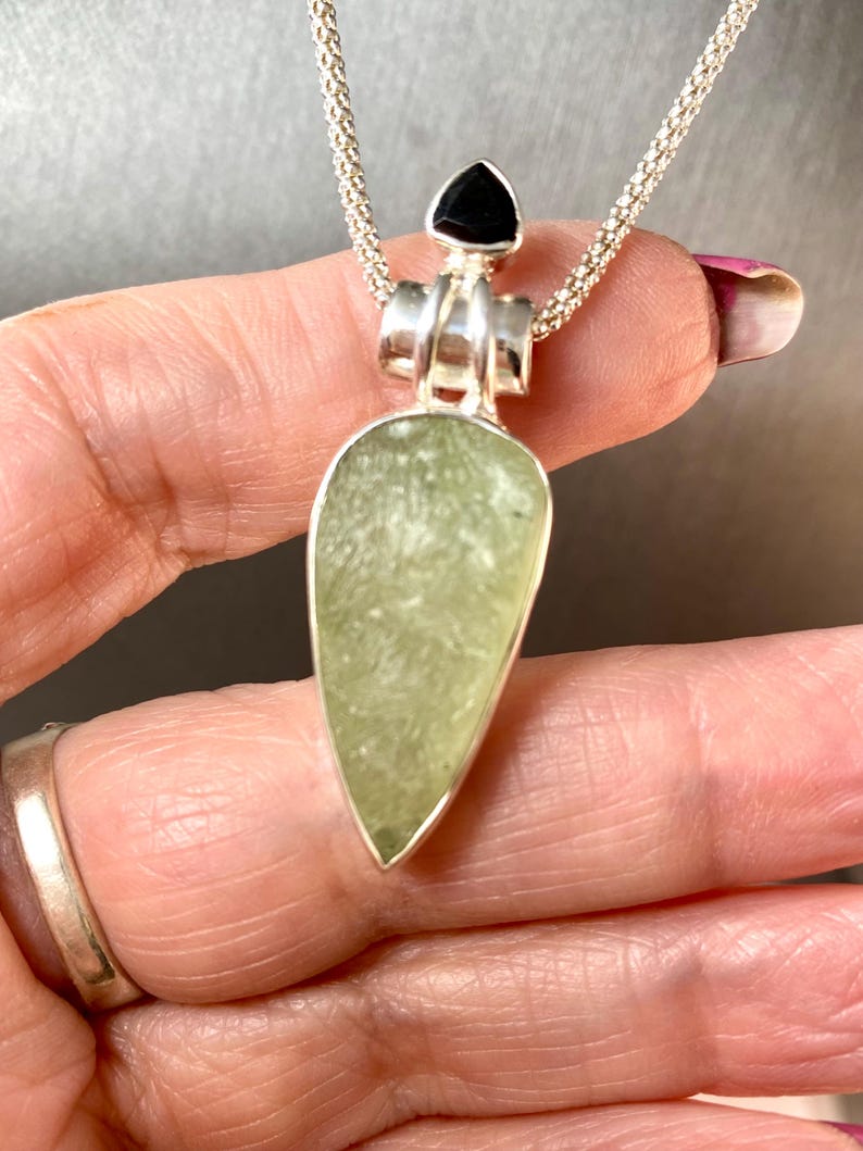 May include: A silver chain necklace with a pendant featuring a light green teardrop-shaped stone and a black triangular stone at the top.