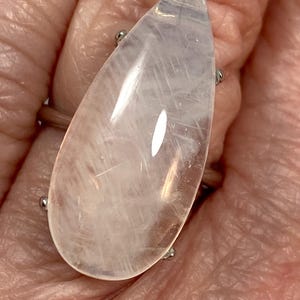 May include: A large, teardrop-shaped ring featuring a translucent, pale pink gemstone. The stone is mounted on a silver-toned band with four small prongs securing the gem. The ring is shown on a person's finger.