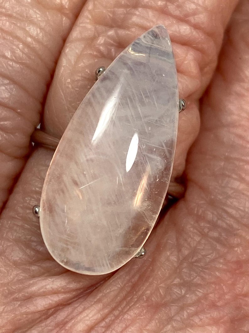 May include: A large, teardrop-shaped ring featuring a translucent, pale pink gemstone. The stone is mounted on a silver-toned band with four small prongs securing the gem. The ring is shown on a person's finger.