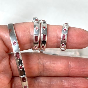 May include: Three silver bangles with pink gemstones. The bangles are stacked on a hand, with the largest bangle on the bottom and the smallest bangle on top.