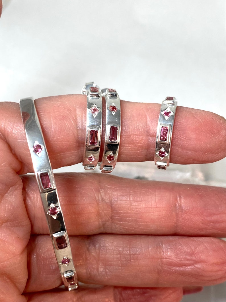 May include: Three silver bangles with pink gemstones. The bangles are stacked on a hand, with the largest bangle on the bottom and the smallest bangle on top.