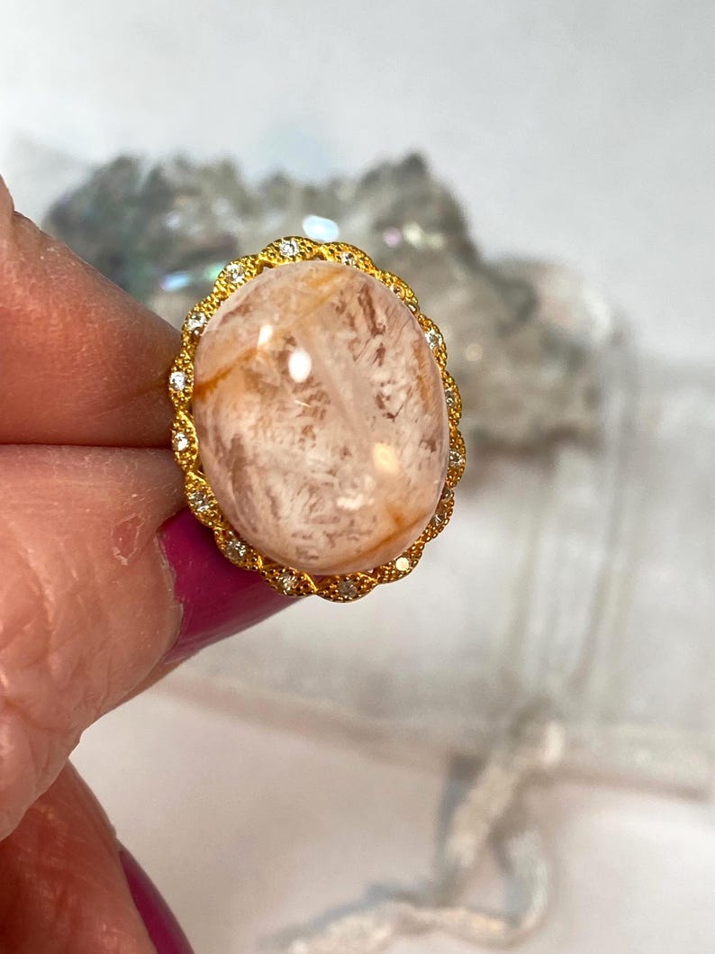 May include: A gold ring featuring a large, oval-shaped, peach-colored gemstone. The gemstone has internal veining and is set in a gold filigree setting with small, clear accent stones. The ring is held up by a person's fingers.