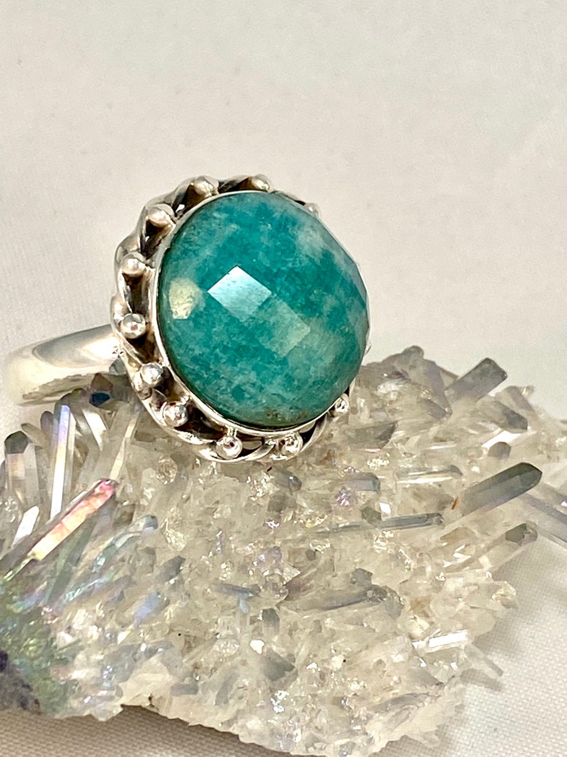 K&ouml;nnte beinhalten: Ein silberner Ring mit einem gro&szlig;en, facettierten, blaugr&uuml;nen Edelstein, der in einer dekorativen, perlenbesetzten Fassung sitzt.