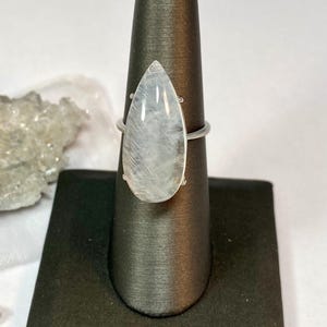 May include: A silver ring featuring a large, pear-shaped, translucent gemstone. The stone has a milky, iridescent quality. The ring is displayed on a black jewelry stand, showcasing its elegant design and craftsmanship.
