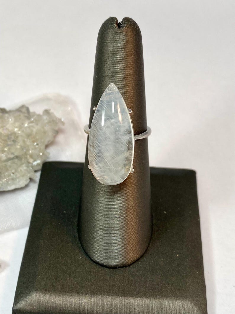 May include: A silver ring featuring a large, pear-shaped, translucent gemstone. The stone has a milky, iridescent quality. The ring is displayed on a black jewelry stand, showcasing its elegant design and craftsmanship.