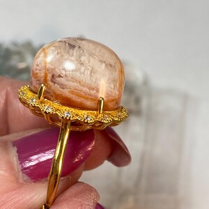 May include: A gold ring featuring a large, orb-shaped gemstone with orange and white striations. The gemstone is set in a gold filigree setting with small, clear accent stones. The ring's band is a simple, polished gold.