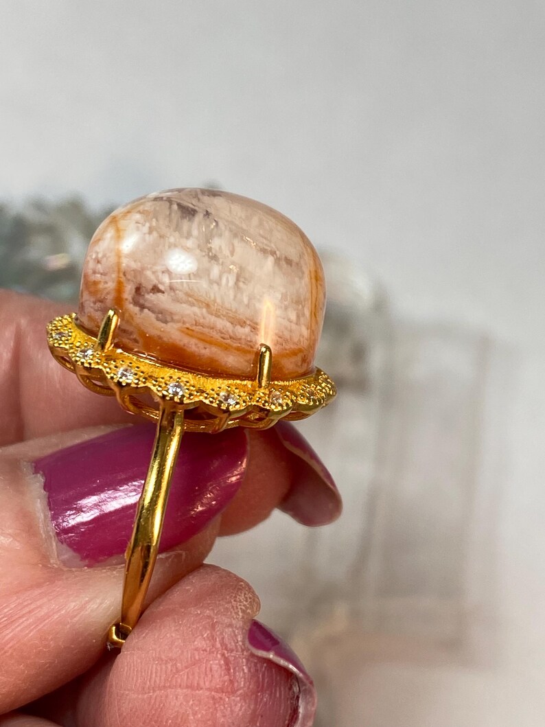 May include: A gold ring featuring a large, orb-shaped gemstone with orange and white striations. The gemstone is set in a gold filigree setting with small, clear accent stones. The ring's band is a simple, polished gold.