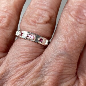 May include: A silver band ring with three pink gemstones. The center stone is rectangular and the two side stones are square.