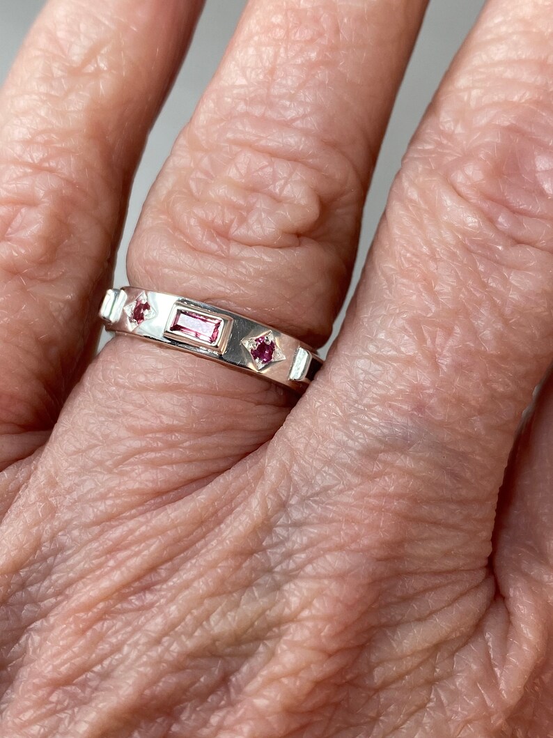 May include: A silver band ring with three pink gemstones. The center stone is rectangular and the two side stones are square.