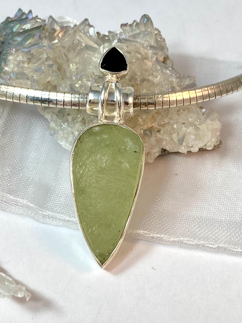 May include: A silver pendant necklace with a light green teardrop-shaped stone and a black triangular stone at the top.