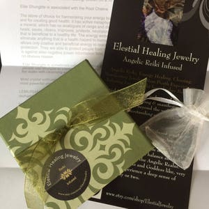 May include: A green gift box with a gold ribbon and a sticker that says "Elestial Healing Jewelry Reiki Infused". The box is sitting on a white surface next to a black card with the text "Elestial Healing Jewelry Angelic Reiki Infused" and a website address.