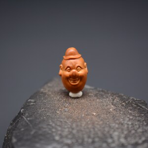 May include: A small, intricately carved wooden figurine of a smiling face with a hat. The figurine is brown and has a detailed expression.