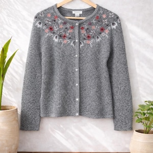 Könnte beinhalten: Grauer Cardigan mit floraler Stickerei am Halsausschnitt. Der langärmlige Pullover hat eine Knopfleiste vorne. Das florale Design umfasst rote, rosa und weiße Blumen. Er hängt an einem Holzkleiderbügel.
