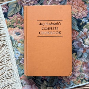May include: A hardcover cookbook with a light orange cover, titled "Amy Vanderbilt's COMPLETE COOKBOOK" in black text. The book is set on a floral patterned fabric with a fringed edge, showcasing a vintage aesthetic.