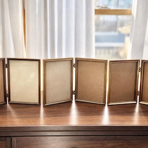 May include: Six hinged, gold-toned picture frames are displayed on a dark wooden surface. The frames are rectangular, with a simple, elegant design. The background features a window with white curtains.