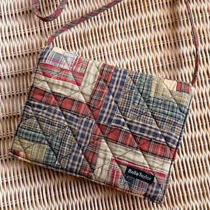 May include: A rectangular quilted crossbody bag featuring a multi-colored plaid pattern in shades of red, blue, green, and tan. The bag has a thin matching plaid strap and a small black fabric label with the white text Bella Taylor attached.