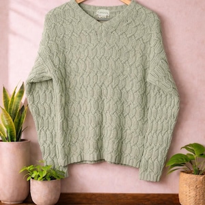 May include: A sage green cable knit sweater with a V-neck, hanging on a wooden hanger. The sweater has a textured pattern and is displayed against a pink background. The word "Lomons" is visible on the label.