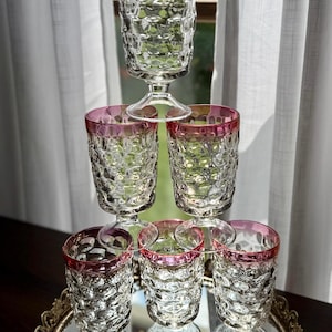 May include: A stack of six clear glass goblets with a textured pattern and pink rims. The goblets are arranged on a mirrored tray, with a seventh goblet balanced on top. The background features a window and white curtains.