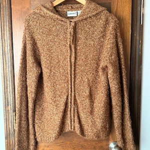 May include: A brown, textured, long-sleeve zip-up hoodie with a knit construction. The garment features a front zipper closure and a label at the neckline that reads Fashion Bug. The material has a flecked appearance with varied brown tones.