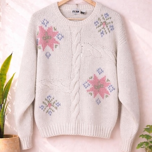 Puede incluir: Suéter color crema con diseños florales rosas, azules y verdes. El suéter tiene cuello redondo, mangas largas y un diseño de punto trenzado. La palabra "ZERO" es visible en la etiqueta.