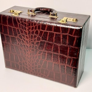 May include: A rectangular dark brown suitcase featuring a textured crocodile skin pattern. The case includes a dark brown top handle and polished gold-tone metal hardware, including two secure latches and hinges along the top edge of the exterior.