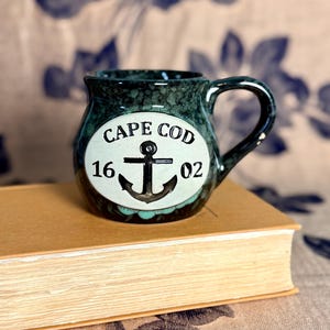 May include: A small, dark green ceramic mug with a handle. The mug features the words "CAPE COD" above an anchor design, with the numbers "16, 02" below. The mug is sitting on a book.