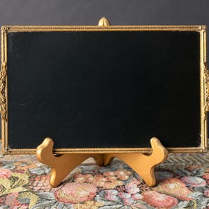May include: A rectangular tray featuring a solid black center panel enclosed by a textured gold-toned metal frame. The frame includes ornate, decorative handles on both sides, characterized by intricate floral and scrollwork detailing.