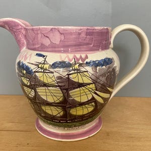 Puede incluir: Una jarra de cerámica con borde y base pintados en color violeta, que presenta una ilustración detallada de un velero en el cuerpo. El barco tiene velas amarillas y aparejos oscuros. El texto "AUSTRALIA CLIPPER SHIP" aparece cerca de la base.