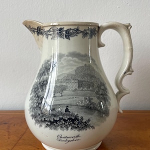 Könnte beinhalten: Ein cremefarbener Keramikkrug mit einem schwarzen Transferdruck von Chatsworth House in Derbyshire. Das Design zeigt eine Landschaftsszene mit Bäumen, Figuren und einem Gebäude, ergänzt durch eine Efeubordüre und den Schriftzug Chatsworth Derbyshire.