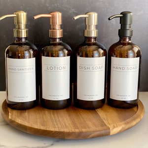 May include: Four amber glass bottles with pump tops and labels. The labels read "Hand Sanitizer", "Lotion", "Dish Soap", and "Hand Soap". The bottles are on a wooden turntable.