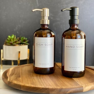May include: Two amber glass soap dispensers with silver and black pumps. The dispensers are labeled "Dish Soap" and "Hand Soap" with the text "The Modern Jar 14 oz" below. The dispensers are on a wooden tray.
