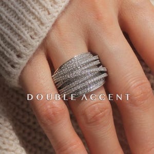 Peut inclure: Une bague argentée ornée de plusieurs rangées de petites pierres scintillantes. La bague est large et couvre une partie importante du doigt. Le texte "DOUBLE ACCENT" est visible en dessous. La bague est posée sur un pull crème.