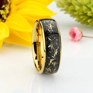 May include: A gold and black tungsten carbide ring with a black inlay and gold flecks. The ring is polished and has a beveled edge.