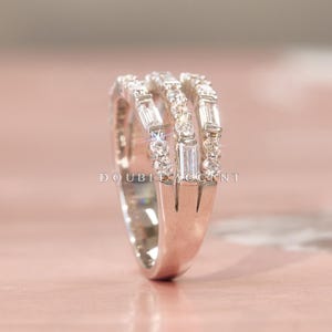 May include: A silver ring with three bands, each adorned with clear, sparkling gemstones. The ring features a mix of baguette and round-cut stones, creating a dazzling display. The text "DOUBLE ACCENT" is visible.
