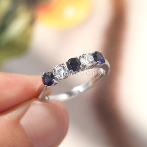 以下が含まれることがあります： 5つの宝石が付いたシルバーリング。宝石は濃い青色と透明で輝く石が交互に並んでいます。リングは手で持ち上げられており、「DOUBLE ACCENT」という文字が一部見えます。