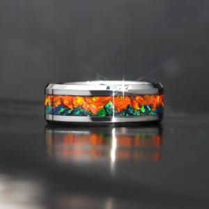 Könnte beinhalten: Ein silberner Ring mit einem Band aus leuchtenden orangefarbenen, grünen und blauen Edelsteinen. Die Edelsteine sind in einem polierten Silberband gefasst, was einen auffälligen Kontrast erzeugt. Das Design des Rings ist modern und auffällig.