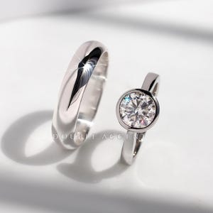 May include: Two silver rings. One is a plain band, the other features a large, round, clear gemstone set in a bezel. The rings are displayed on a white surface. The text "DOUBLE ACCENT" is visible.